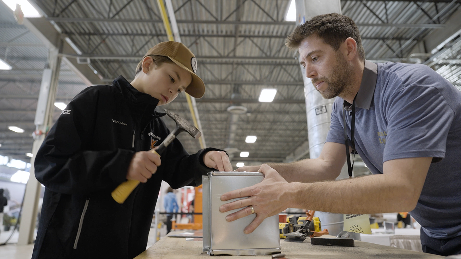 A student at career fair is using a hammer on a metal while the technician assisting.